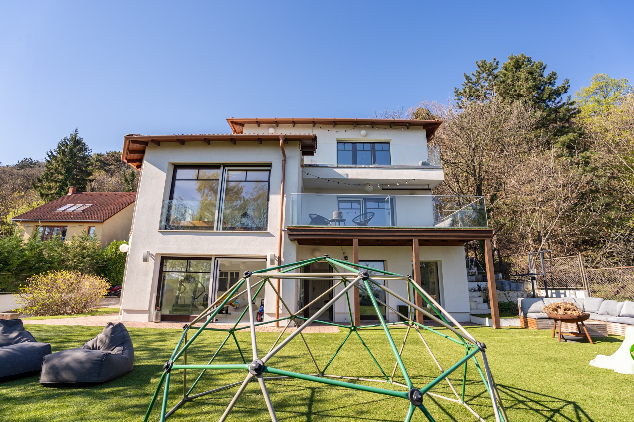 Egy modern családi ház hátsó homlokzata látható egy rendezett, zöld gyepes udvarral. Az ingatlanhoz egy tágas erkély és alsó terasz is tartozik, ahonnan közvetlenül megközelíthető a kert. Az udvaron egy játszótéri mászóka és kényelmes kültéri bútorok állnak. Napos, tiszta az idő, zöldellő fák és bokrok veszik körül a telket.