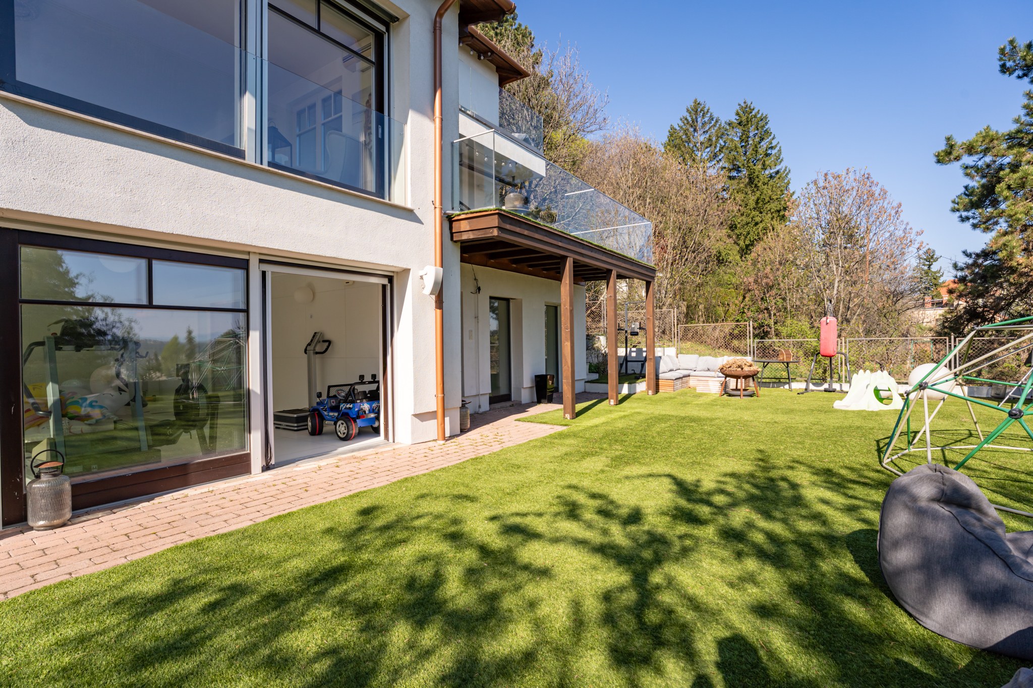 The image shows the sun-drenched garden of a modern family home with artificial grass, a terrace area and various leisure equipment. Through the open glass doors, an interior space with exercise machines and toys is visible.