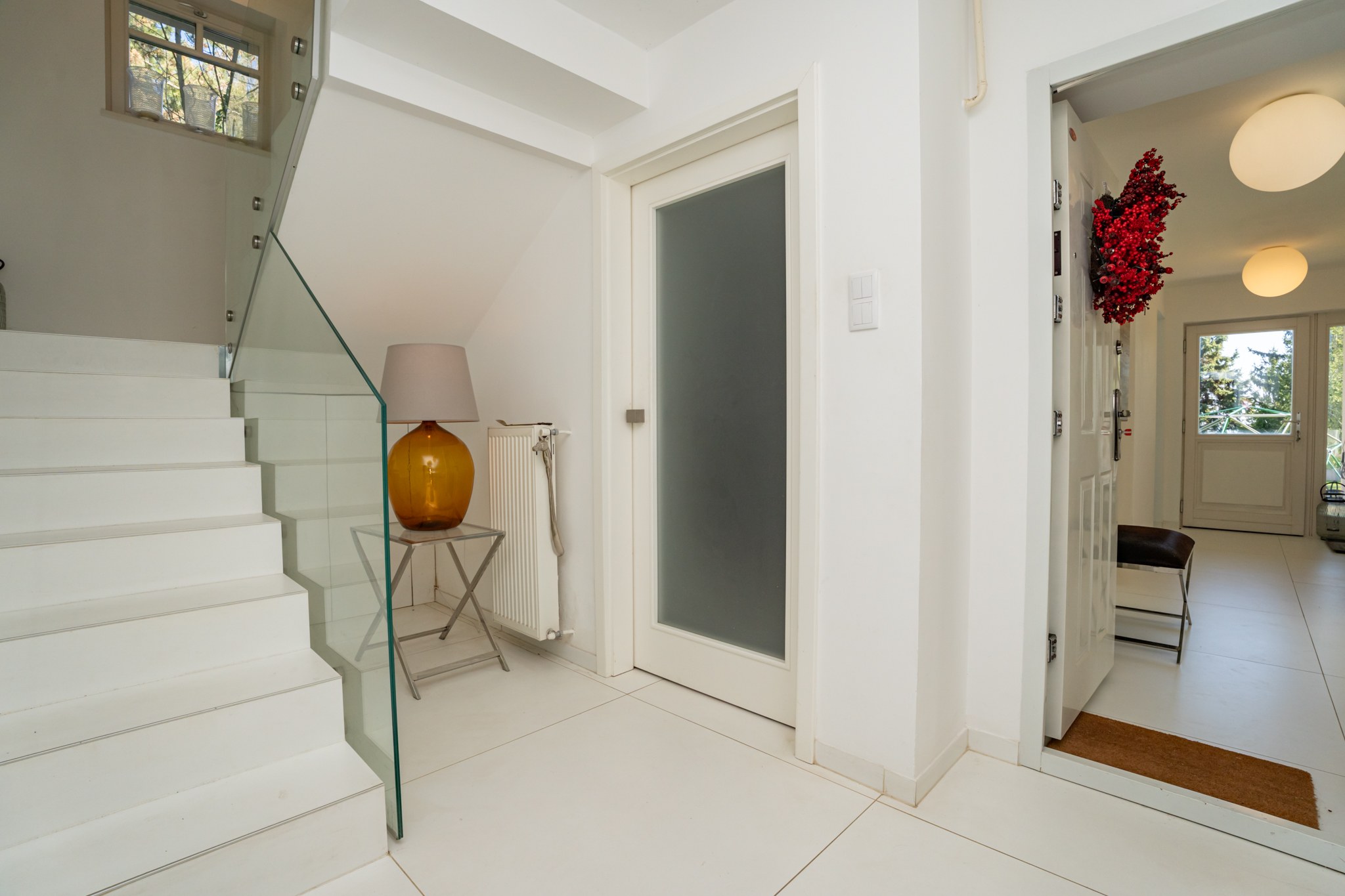 A bright, modern staircase landing and hallway inside the property. Characterised by white tiled flooring, a contemporary white staircase and a glass balustrade. One door stands open with a view towards an exterior area or terrace.