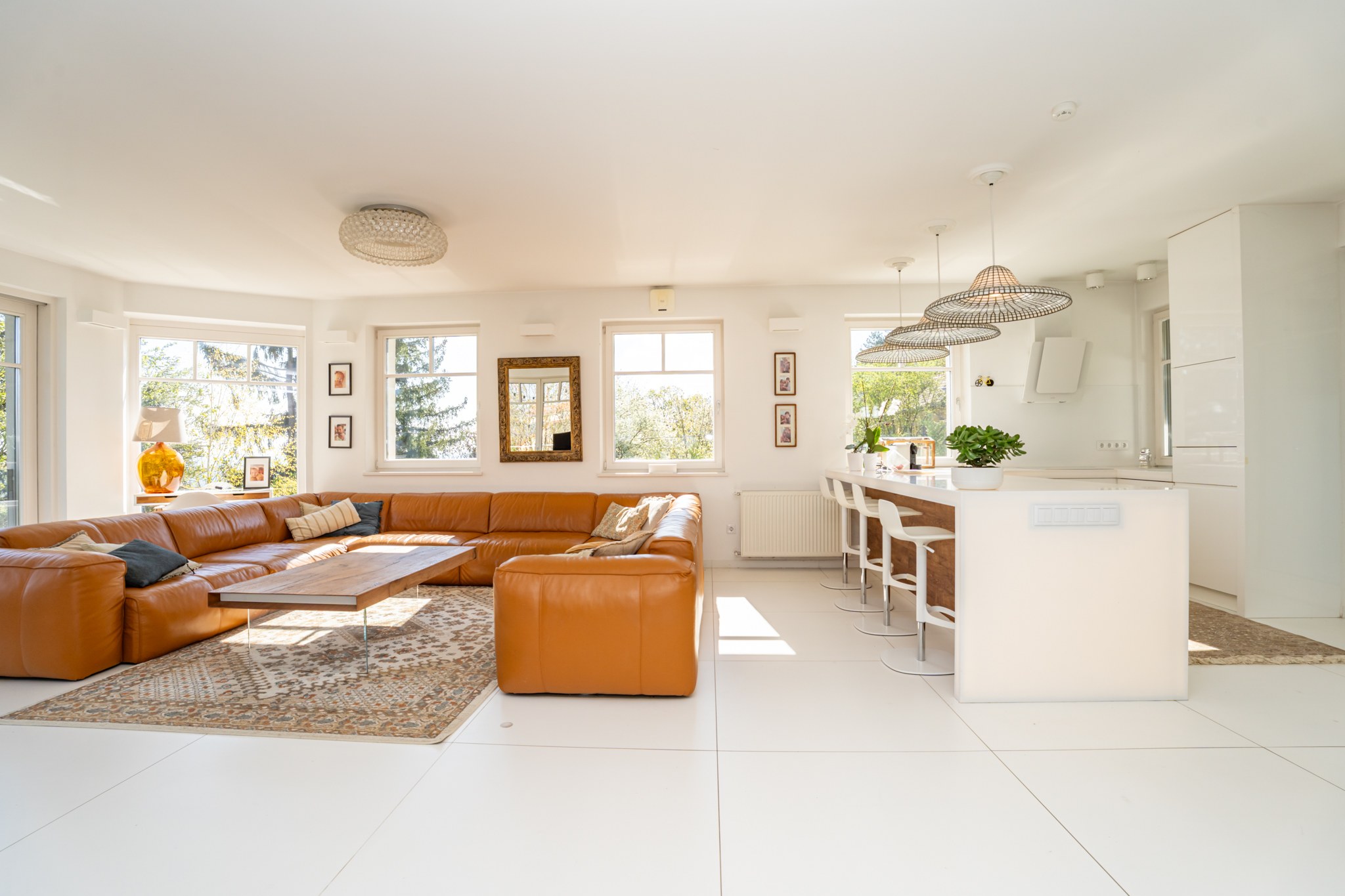A modern, open-plan living-kitchen area with a brown leather corner sofa and a sleek kitchen island. The space is exceptionally bright thanks to numerous large windows, with white floors and walls throughout.