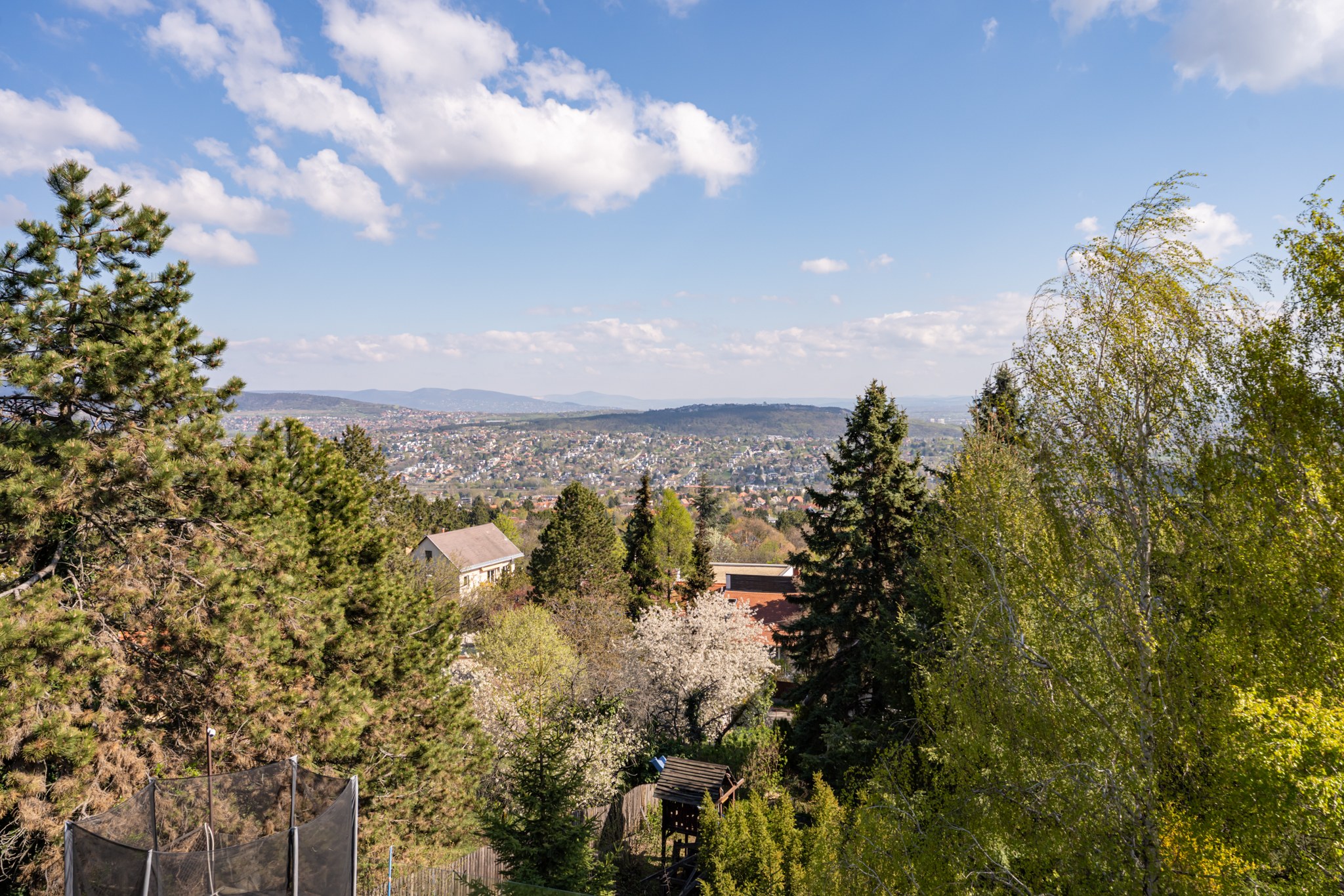 Panorámás kilátás egy domboldali, sűrűn beépített lakóövezetre, távoli hegyekkel és tiszta kék éggel, fehér felhőkkel. Az előtérben dús zöld növényzet, néhány ház teteje és egy trambulin látható.