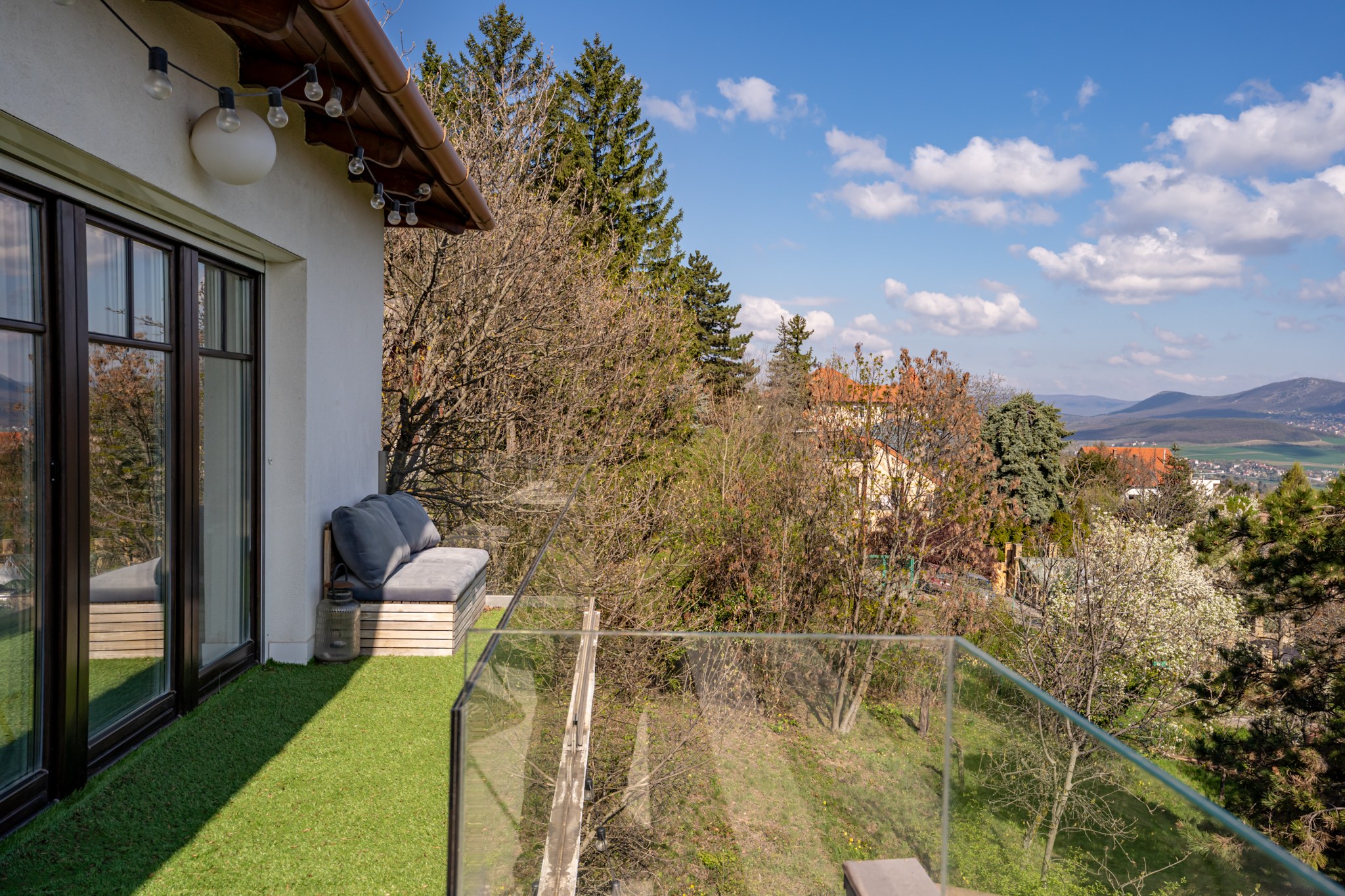 Ez egy modern erkély vagy terasz, műfű borítással, kényelmes kerti ülőpaddal és üveg korláttal. Fák övezik, és lenyűgöző panoráma nyílik a környező dombokra és a tájra.
